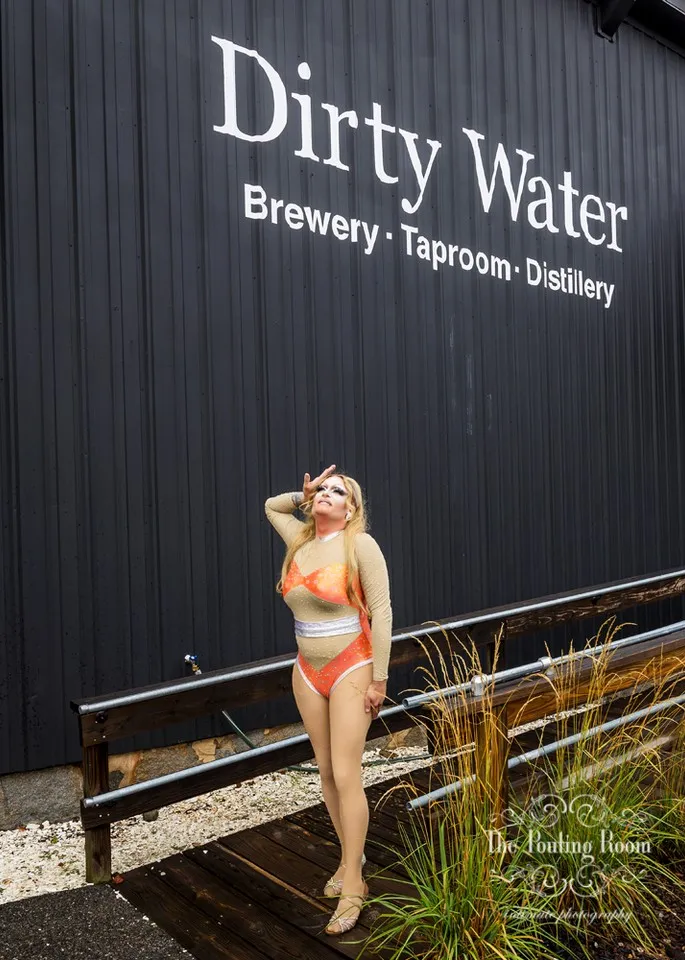 A drag performer posing outside the Dirty Water Distillery sign